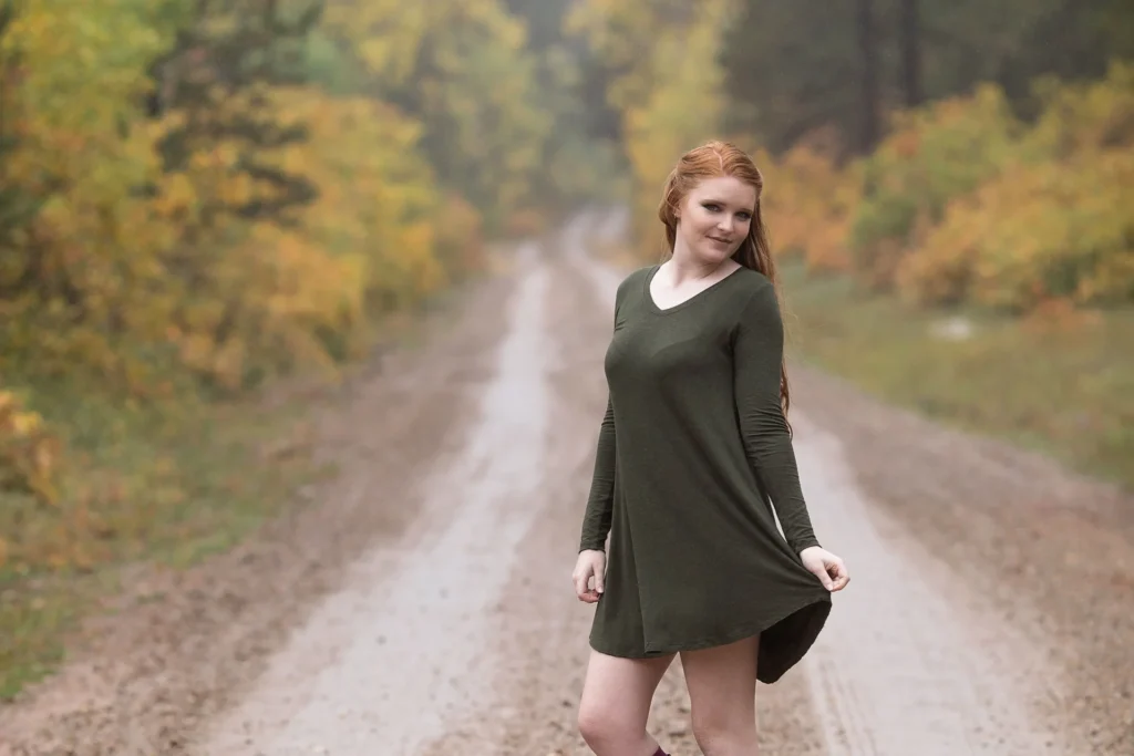 Młoda kobieta w eleganckiej sukience pozuje pewnie na polnej drodze wśród jesiennych liści.