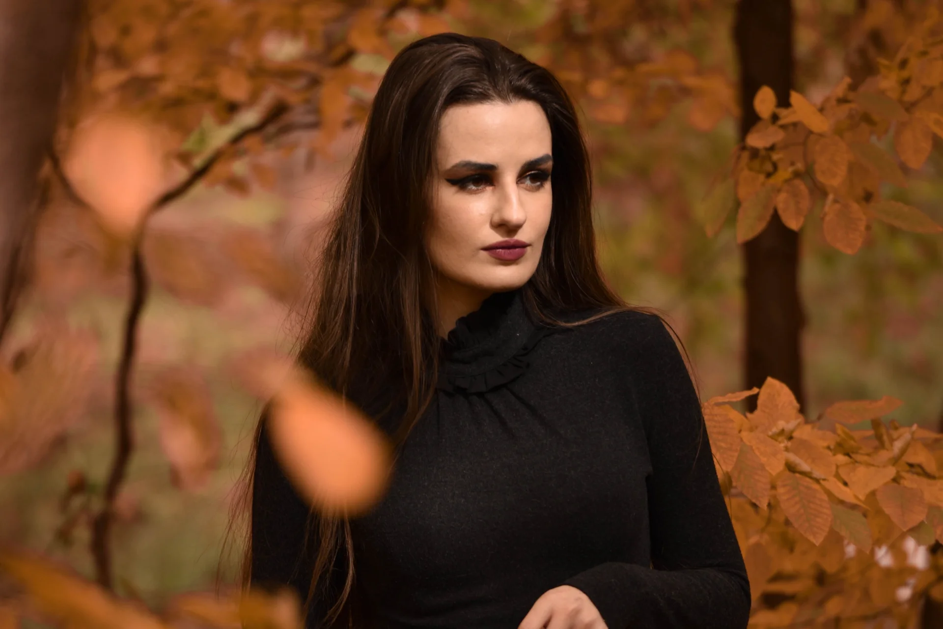 Młoda kobieta w eleganckiej czarnej sukience pozuje w jesiennym lesie, otoczona liśćmi.